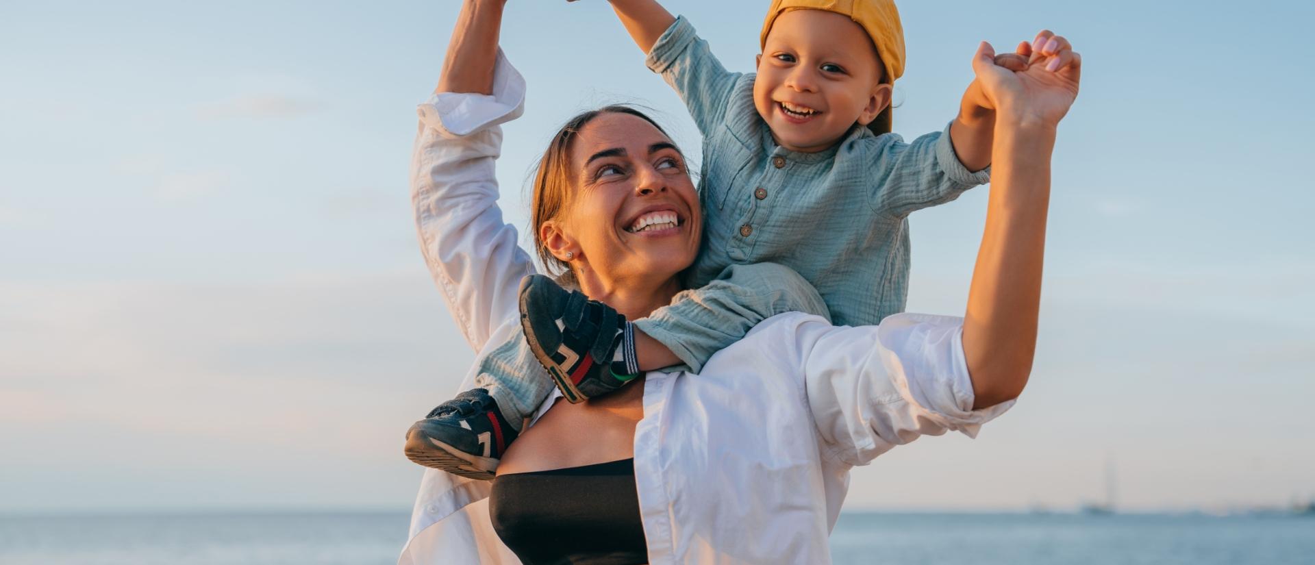 Madre felice con bambino sulle spalle, sorridono insieme al tramonto sulla spiaggia.
