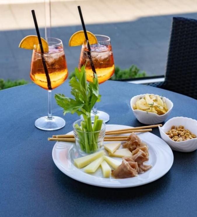 Aperitivo con Spritz, formaggi, prosciutto, sedano e snack su un tavolo all'aperto.