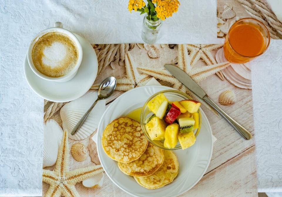 Colazione con pancake, macedonia, cappuccino e succo su tovaglietta decorata con conchiglie.